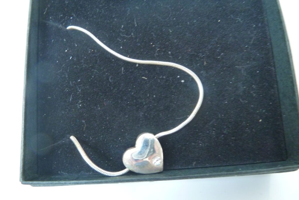 Silver-tone snake chain necklace with a small, smooth heart pendant featuring a clear crystal accent, displayed in a black velvet box